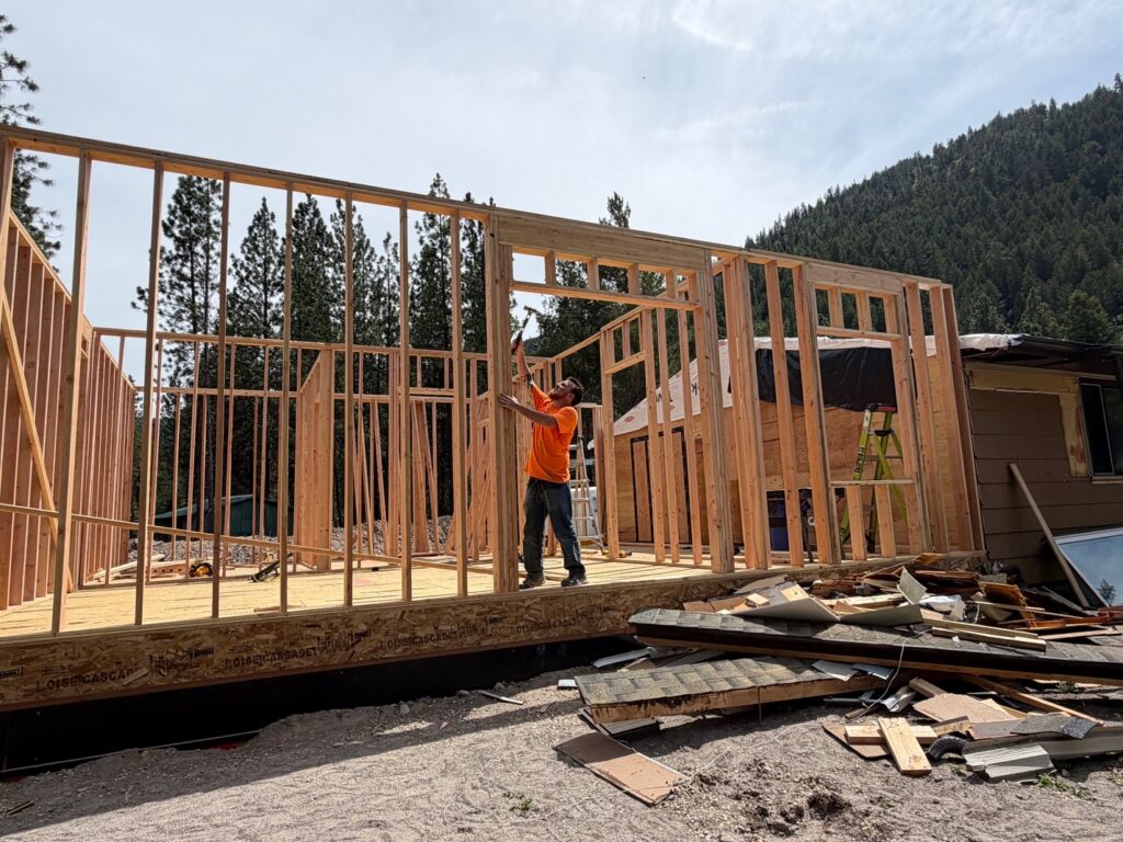 A worker in an orange shirt uses a hammer while framing the wooden wall of a house under construction, with forested mountains in the background and a pile of construction debris nearby.