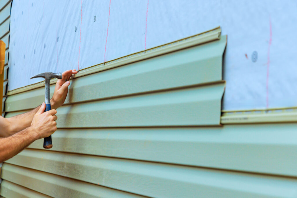 Worker's hands using a hammer to install green vinyl siding panels over house wrap on an exterior wall