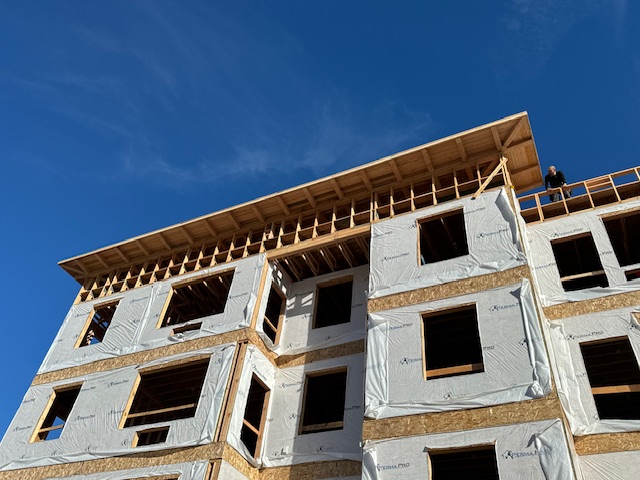Multi-story apartment building framed by Advanced Custom Builders with a worker on the upper level under a clear Montana sky