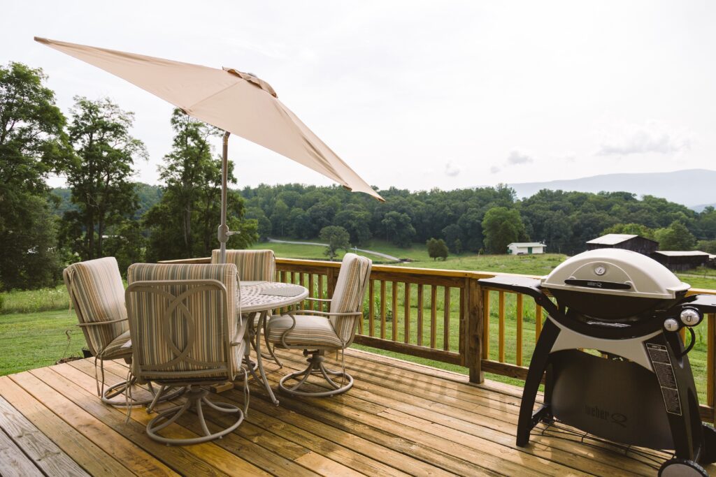 A wooden deck with striped cushioned patio furniture, a beige umbrella, and a Weber grill overlooking lush green countryside and rolling hills.