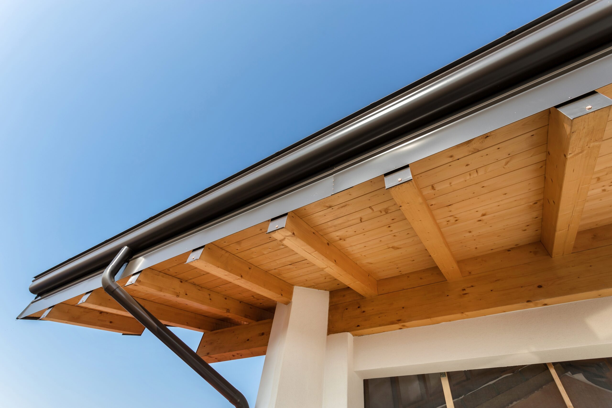 Dark metal gutter and downspout along a wooden roof eave with exposed timber beams against a clear blue sky